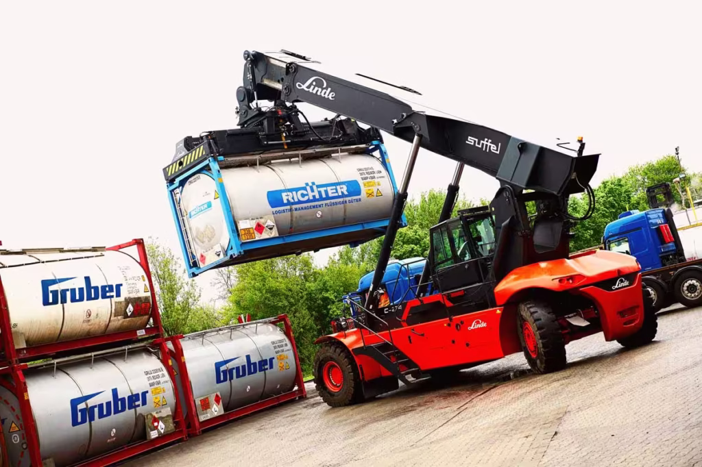 Ein großer rot-schwarzer Gabelstapler hebt einen chemischen Metalltank mit der Aufschrift "Richter" neben gestapelten Behältern mit der Aufschrift "Gruber" in einem Industriegebiet an. Im Hintergrund sind Bäume und ein blauer Lastwagen zu sehen.