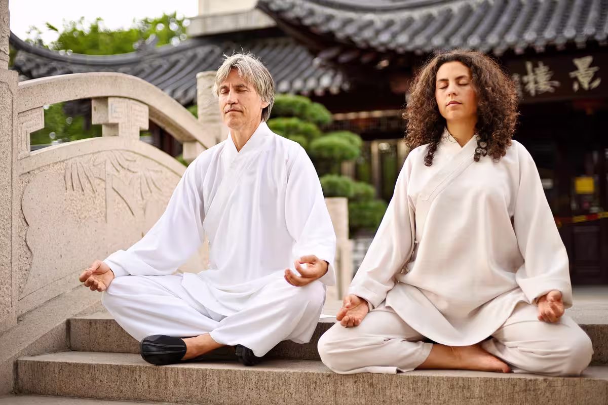 Zwei Personen in weißer traditioneller Kleidung sitzen im Schneidersitz auf Steinstufen und meditieren mit geschlossenen Augen vor einem verzierten Gebäude mit geschwungenem Dach und dekorativen Elementen. Business-Fotografie Mannheim, Heidelberg und Ludwigshafen