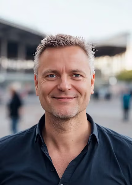 Axel Rosenberg, ein Mann mit kurzen, hellen Haaren und einem dunklen Button-up-Hemd, lächelt, während er im Freien vor einer Stadtkulisse mit unscharfen Gebäuden und Menschen im Hintergrund steht. Fotoherold - Fotografenteam Mannheim-Heidelberg
