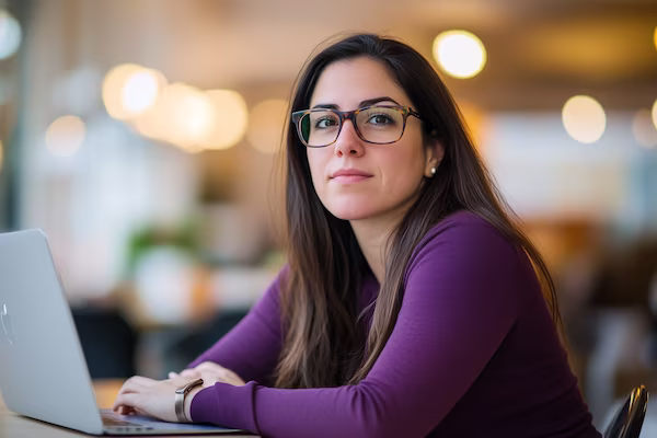 Eine Frau mit langen braunen Haaren und Brille sitzt an einem Tisch mit einem Laptop und schaut in die Kamera. Sie trägt ein lila Oberteil und befindet sich in einem hell beleuchteten Innenraum.