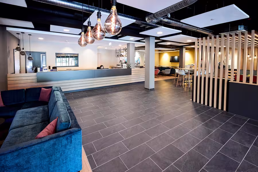 Eine moderne Hotellobby mit dunklem Fliesenboden, einer blauen Couch, Hängelampen, einem hölzernen Empfangstresen und einer Trennwand mit vertikalen Holzlatten. Im Hintergrund sind Tische und Stühle zu sehen.