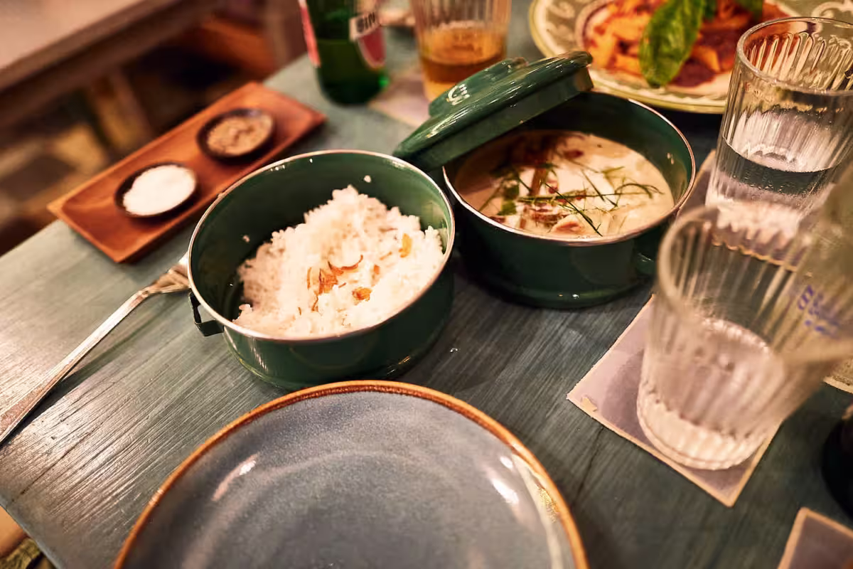 Auf einem im Stil der modernen Gastrofotografie gedeckten Tisch stehen eine Schüssel mit weißem Reis, eine cremige Suppe mit Kräutergarantie, ein leerer Teller, zwei Gläser und ein Holztablett mit Salz und Pfeffer. Im Hintergrund sind eine Flasche und ein weiteres Gericht zu sehen. Fotoherold - Fotografenteam Mannheim-Heidelberg