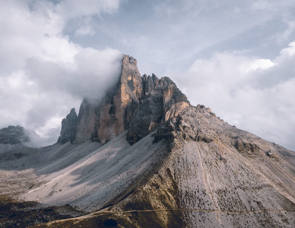 Felsige Berggipfel erheben sich steil über eine abschüssige, karge Landschaft. Der höchste Gipfel ist teilweise bewölkt, und der Himmel ist bedeckt. An den unteren Hängen ist spärliche Vegetation zu sehen.