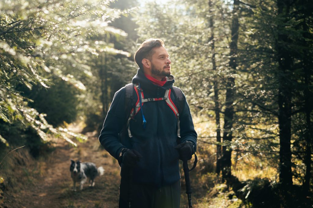 Ein Mann mit Rucksack und Handschuhen steht mit Wanderstöcken in der Hand auf einem Waldweg. Das Sonnenlicht fällt durch die Bäume. Ein Hund läuft hinter ihm auf dem Weg.